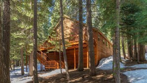 A log cabin with a wooden deck and staircase, surrounded by tall trees and snow on the ground. - Everything You Need in a Mountain Cabin (Truckee)