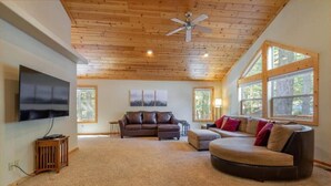 A spacious living room with a high ceiling, wooden beams, a ceiling fan, a flat-screen TV mounted on the wall, a brown sectional sofa, a beige armchair, and large windows providing natural light. - Everything You Need in a Mountain Cabin (Truckee)