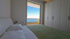 A bedroom with a bed, a nightstand, and a view of the sea through an open door. - Planet Apartments (Sarandë)