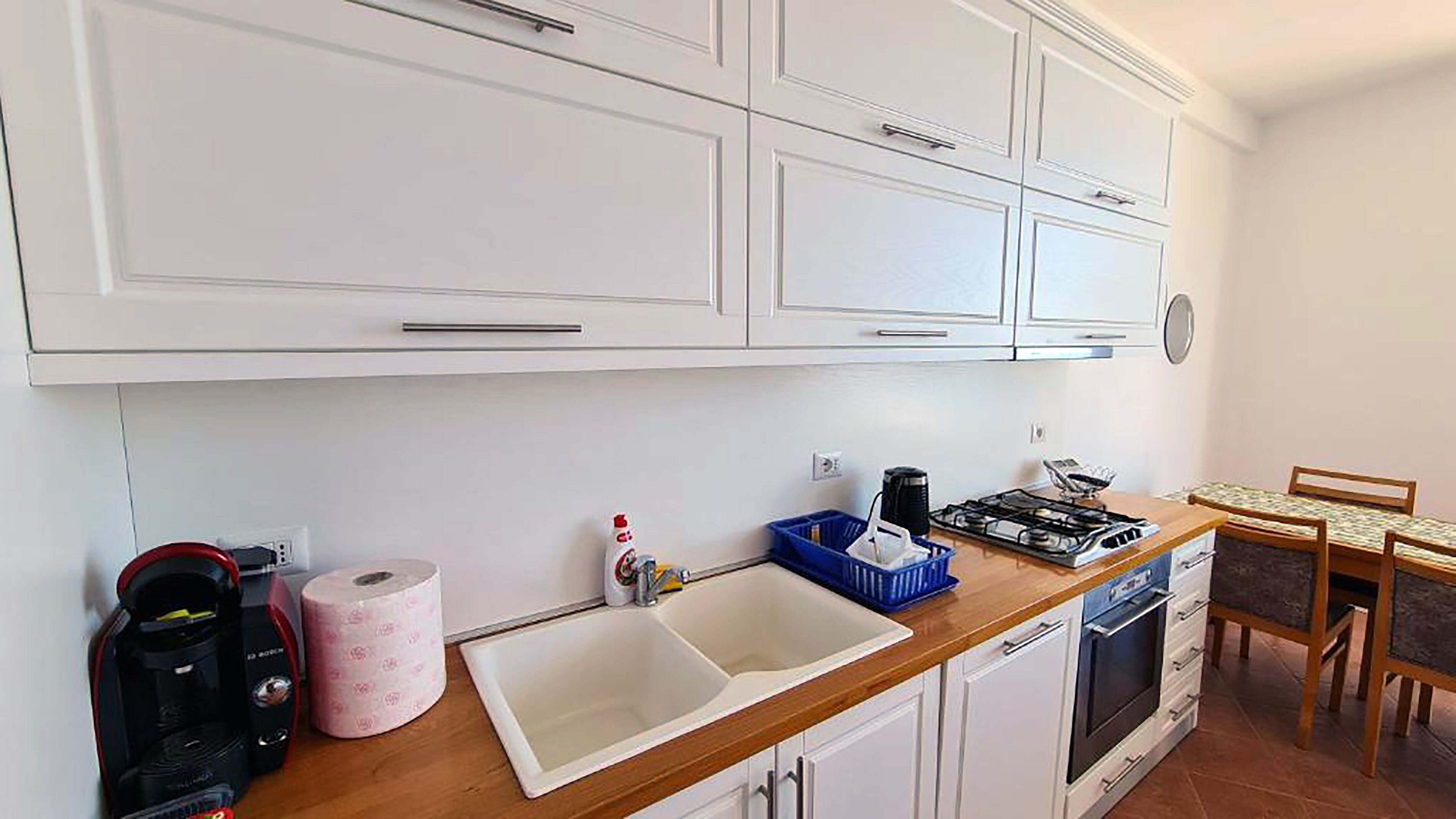 Une cuisine avec des meubles blancs, un évier, une cuisinière et un coin repas avec des chaises.