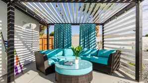Un área de estar al aire libre con un sofá y una mesa redonda, bajo una pérgola con tela a rayas.