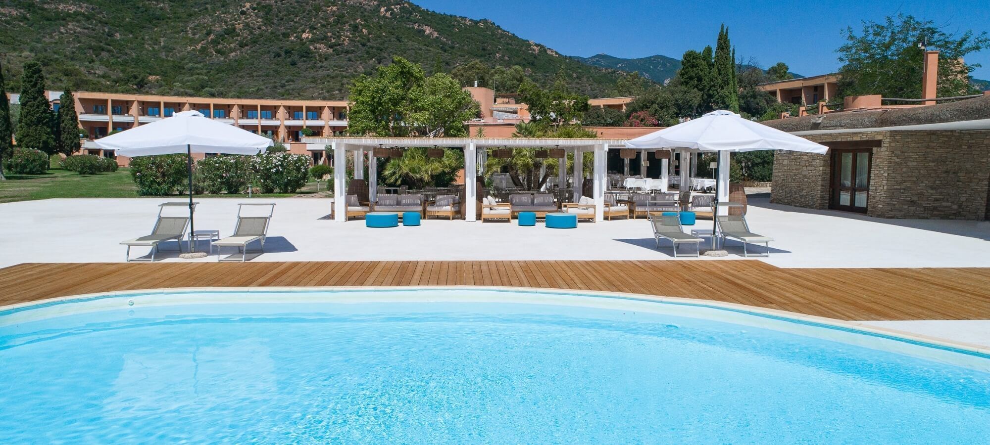 A pool area with lounge chairs and umbrellas, surrounded by buildings and a mountainous backdrop.