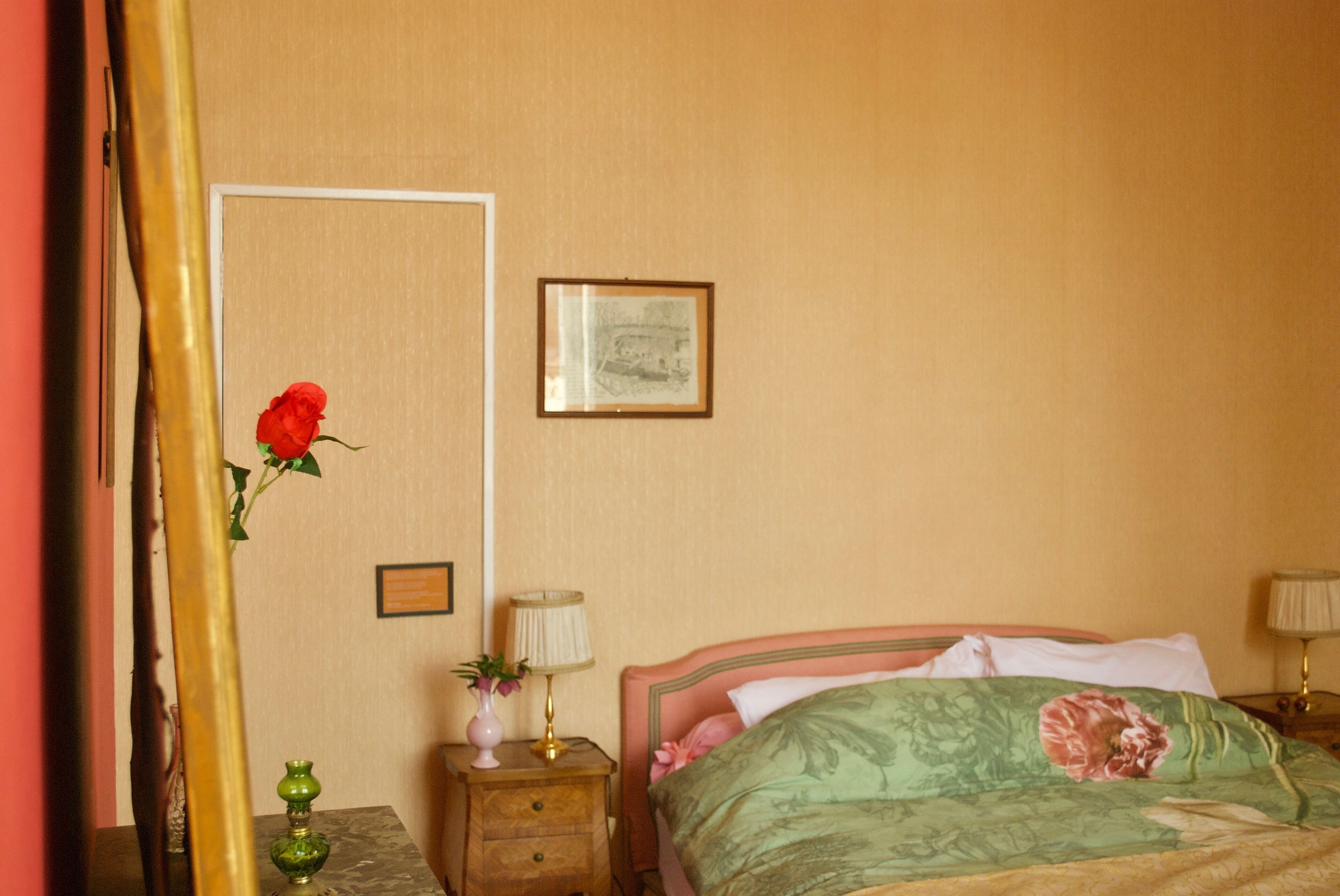 A bedroom with a bed, a nightstand, a lamp, a framed picture, and a vase with a flower.