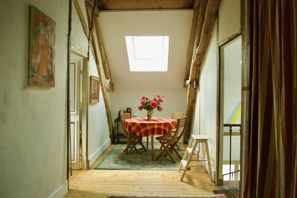Una habitación estrecha con suelo de madera, una mesa con mantel a cuadros y un jarrón con flores.