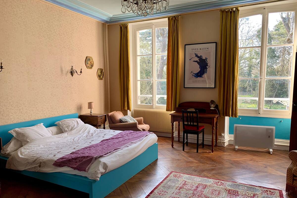 Un dormitorio con una cama azul, un escritorio, una silla, un radiador y un ventanal grande con cortinas.