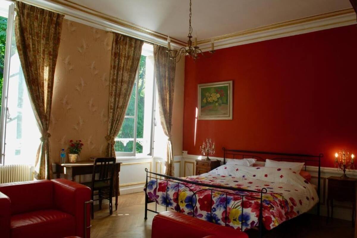 Un dormitorio con cama, pared roja, ventana con cortinas, escritorio y silla.