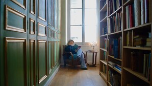 A room with a large window, a wooden floor, a chair, and a bookshelf filled with books.