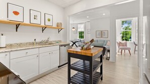 A modern kitchen with white cabinets, a granite countertop, and a wooden table with a black metal shelf. There is a dining area with a light blue sofa and a pink chair. - Physician`s Loft in the heart of downtown with 2 Free Parking Spots (Bar Harbor)