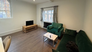 Ein Wohnzimmer mit einem grünen Sofa, einem Holztisch, einem Fernseher auf einem Schrank und einem Fenster mit gemusterten Vorhängen.