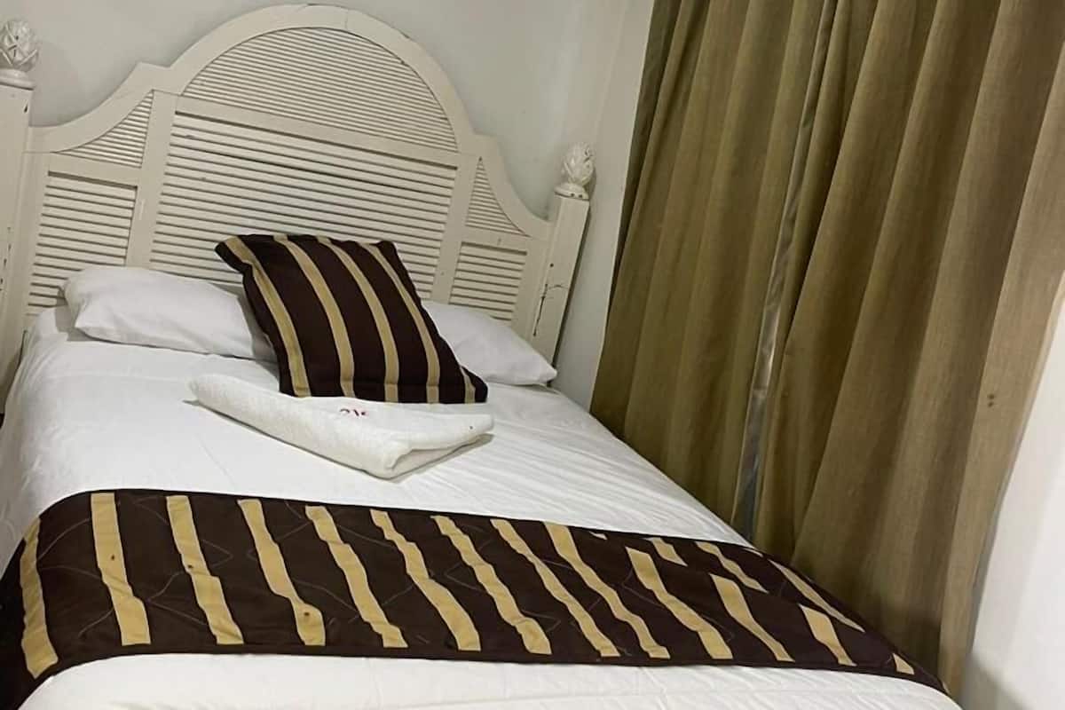 Habitación de hotel con cama, cabecera blanca, equipo de aire acondicionado y cortinas.