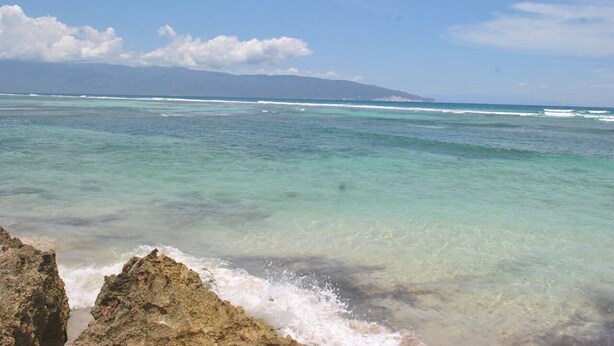 A coastal view with clear turquoise waters, rocky shoreline, and distant mountains.
