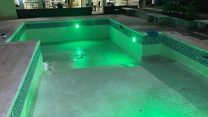 A swimming pool with green lighting, surrounded by a tiled deck and outdoor furniture.