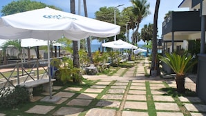 A paved pathway lined with white umbrellas, palm trees, and a building with a balcony overlooking the ocean.
