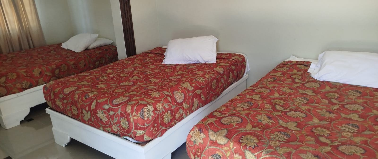Une chambre d’hôtel avec trois lits simples, chacun recouvert d’une courtepointe à motifs floraux rouges et agrémenté d’oreillers blancs.
