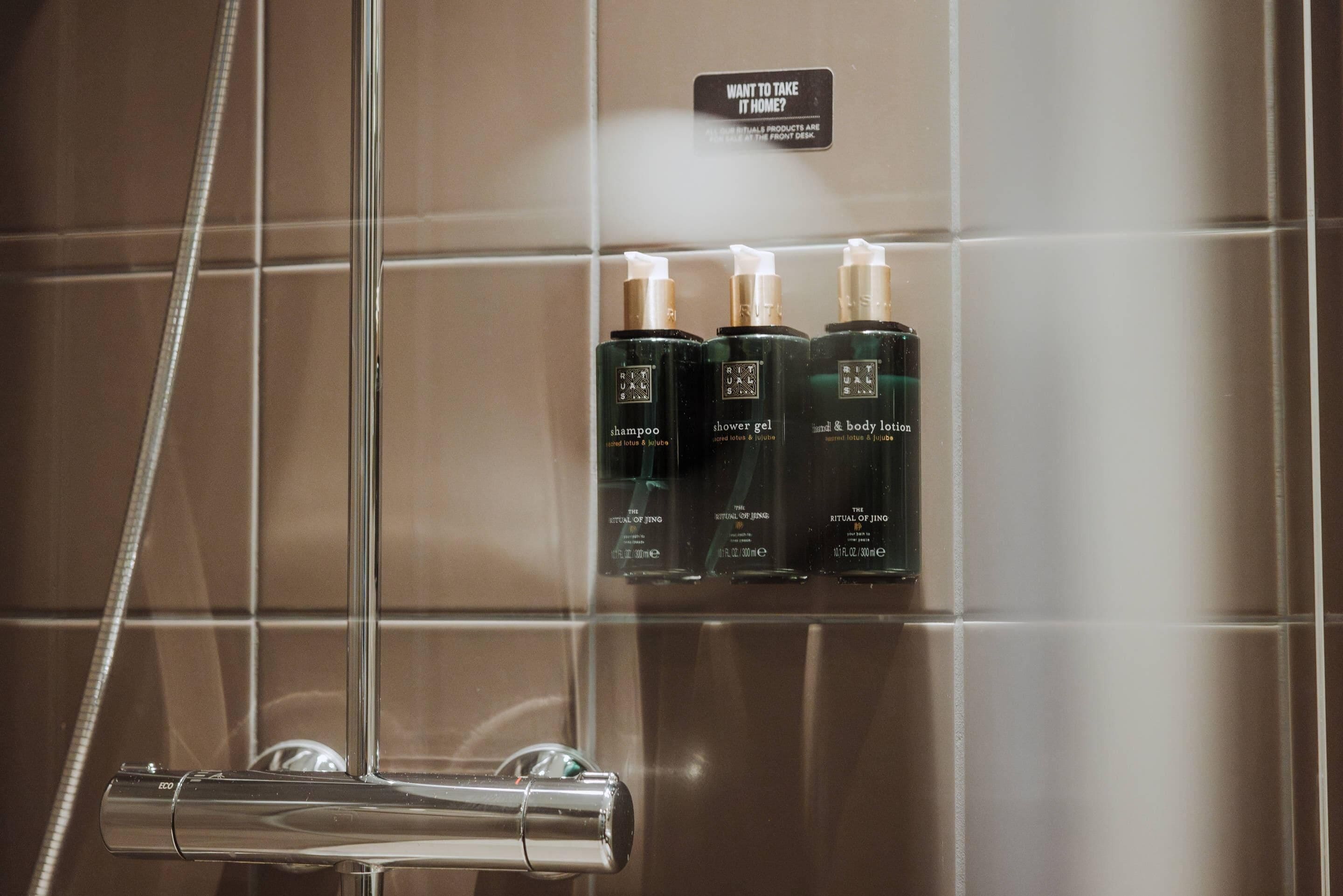 A shower area with a glass door, a showerhead, and three bottles of shampoo, shower gel, and body lotion on the wall.