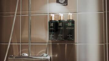 A shower area with a glass door, a showerhead, and three bottles of shampoo, shower gel, and body lotion on the wall.