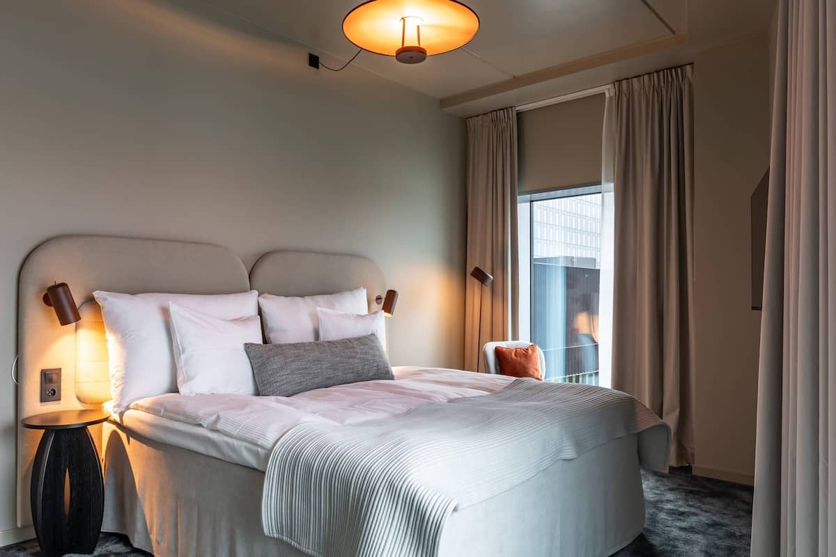 Ein Hotelzimmer mit einem großen Bett, einem Nachttisch mit Lampe, einem Fenster mit Vorhängen und Blick auf ein Gebäude.