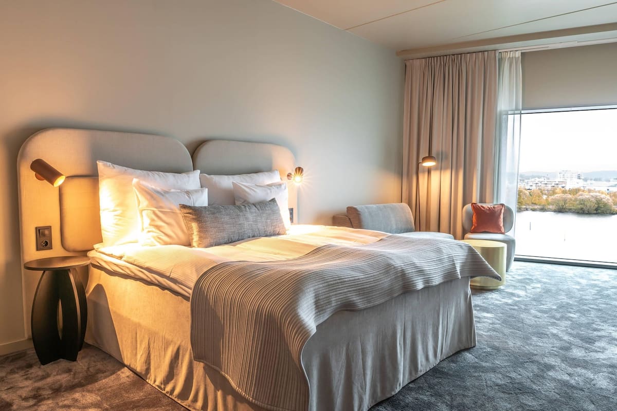 Ein Hotelzimmer mit einem großen Bett, Nachttischlampen, einem Sessel und Blick auf die Stadt.