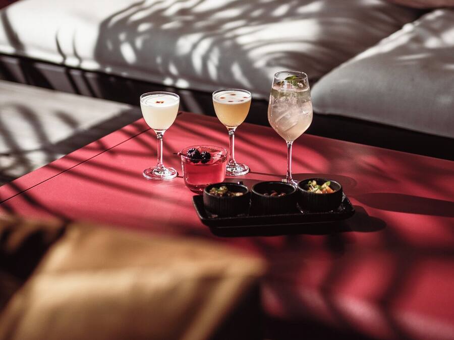Una cama con cabecera roja, un almohadón blanco y un almohadón marrón. Hay tres vasos con bebidas sobre una mesa roja frente a la cama.