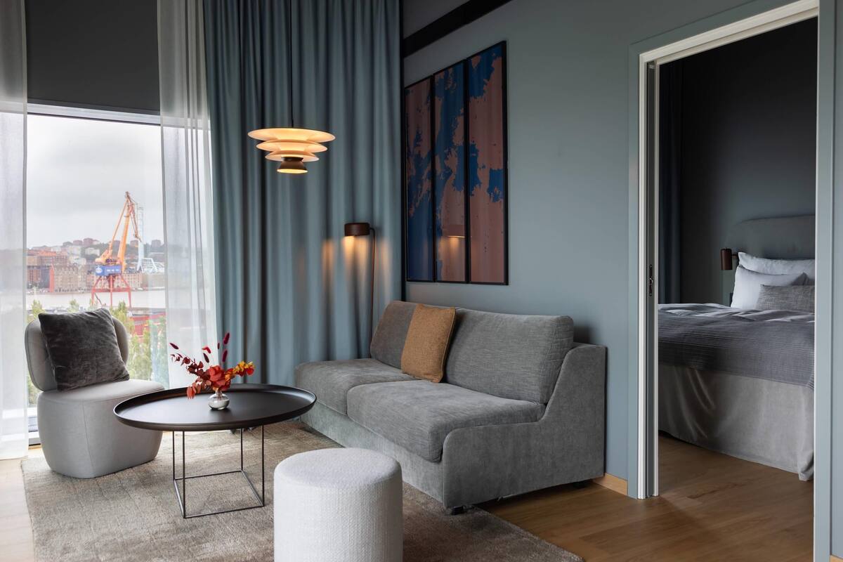 Ein modernes Hotelzimmer mit grauem Sofa, einem runden Couchtisch, einem kleinen Beistelltisch und einem Bett mit grauem Kopfteil.