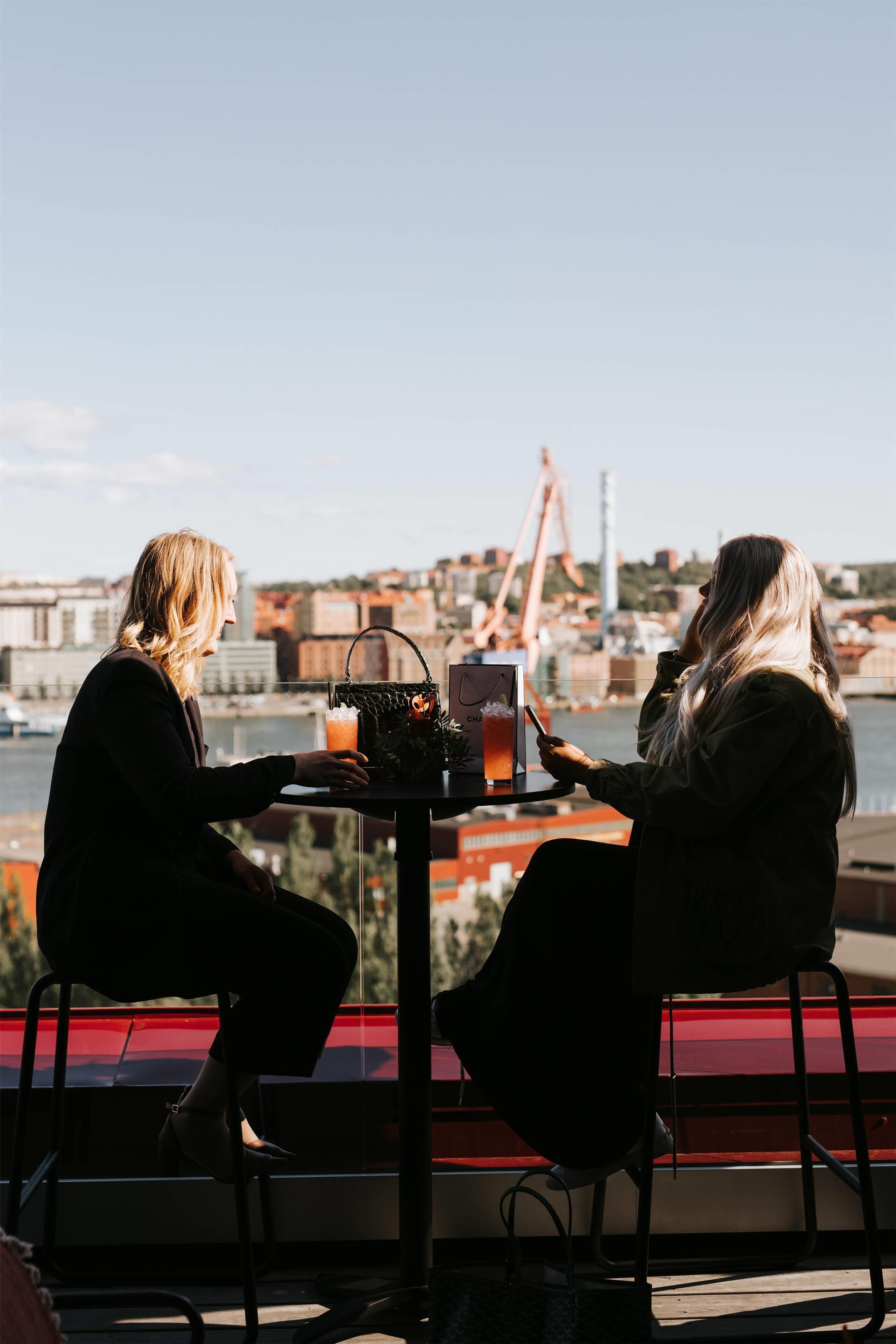 Two people seated at a table with a drink and a handbag, overlooking a waterfront with buildings and a crane in the background.