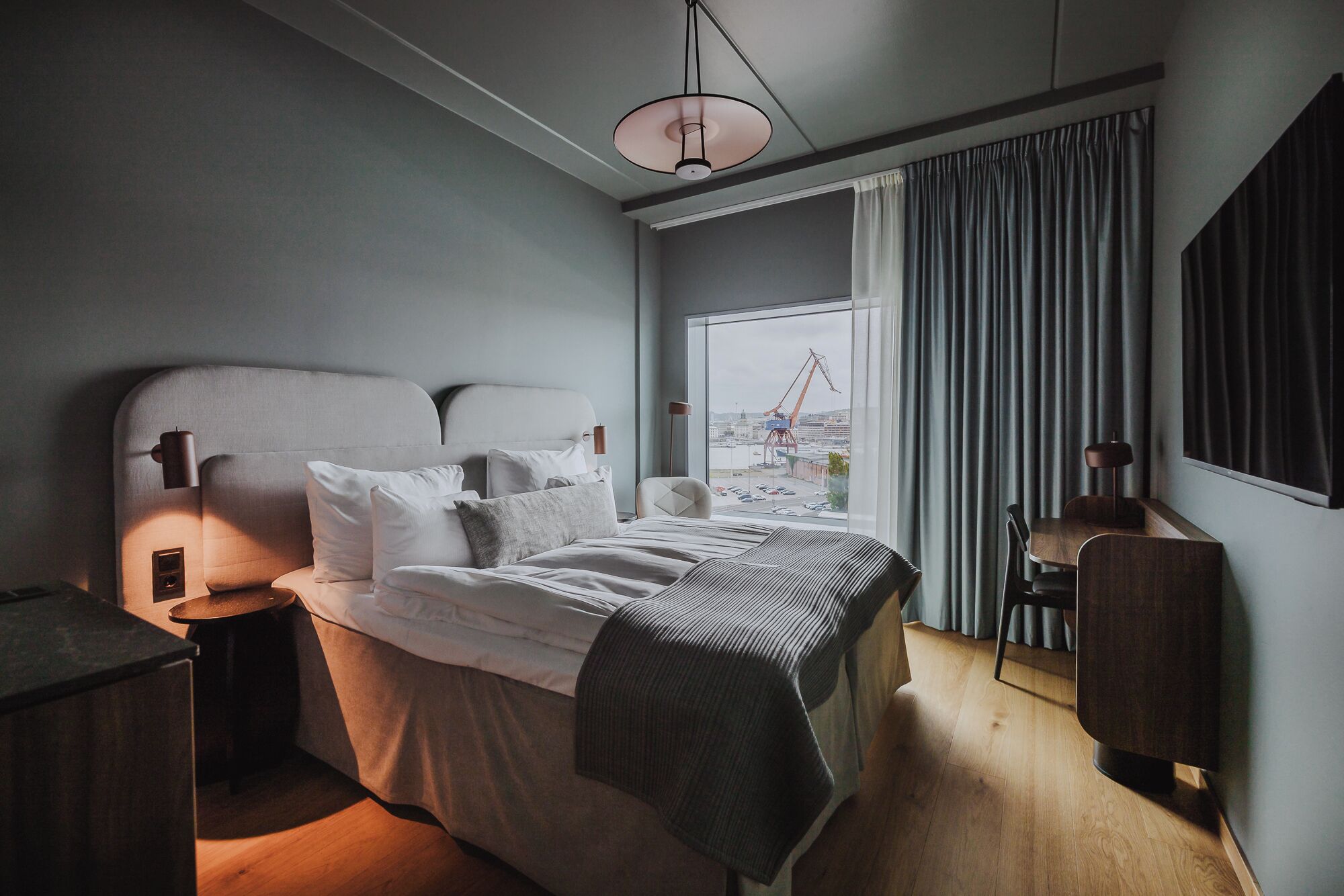 A modern bedroom with a large bed, bedside lamps, a view of a crane through the window, and a wooden desk with a chair.