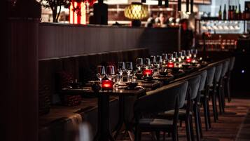 Restaurante com iluminação suave, mesa de jantar comprida para vários convidados, luminárias pendentes no teto e um bar ao fundo.