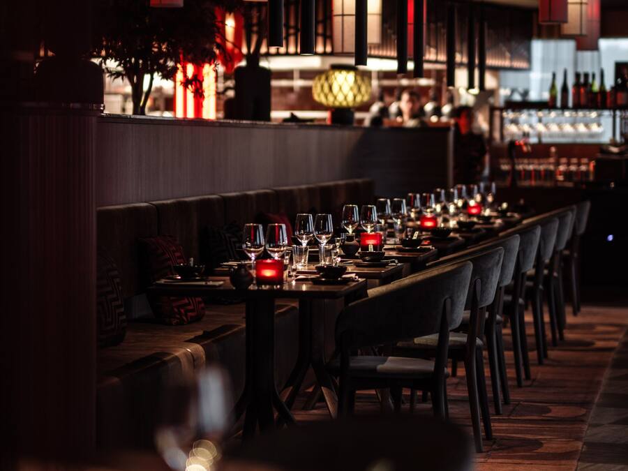 Restaurante com iluminação suave, mesa de jantar comprida para vários convidados, luminárias pendentes no teto e um bar ao fundo.