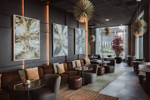 A modern lounge area with a sofa, armchairs, and tables, featuring abstract wall art and pendant lighting.