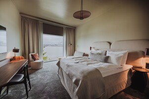 Quarto de hotel com uma cama grande, uma escrivaninha, uma cadeira, um abajur e vista para um corpo d'água pela janela.