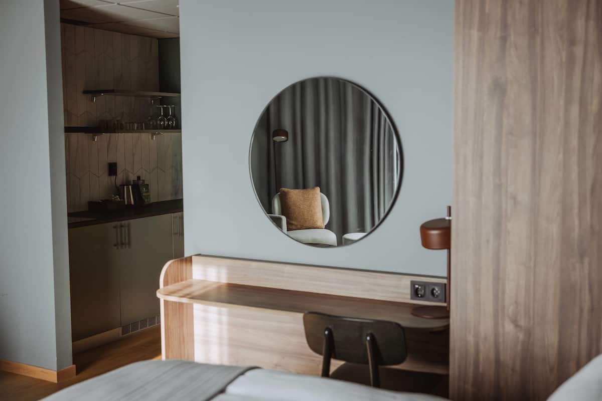 Ein modernes Hotelzimmer mit Schreibtisch aus Holz, einem runden Spiegel, einem Stuhl und einer kleinen Küche im Hintergrund.
