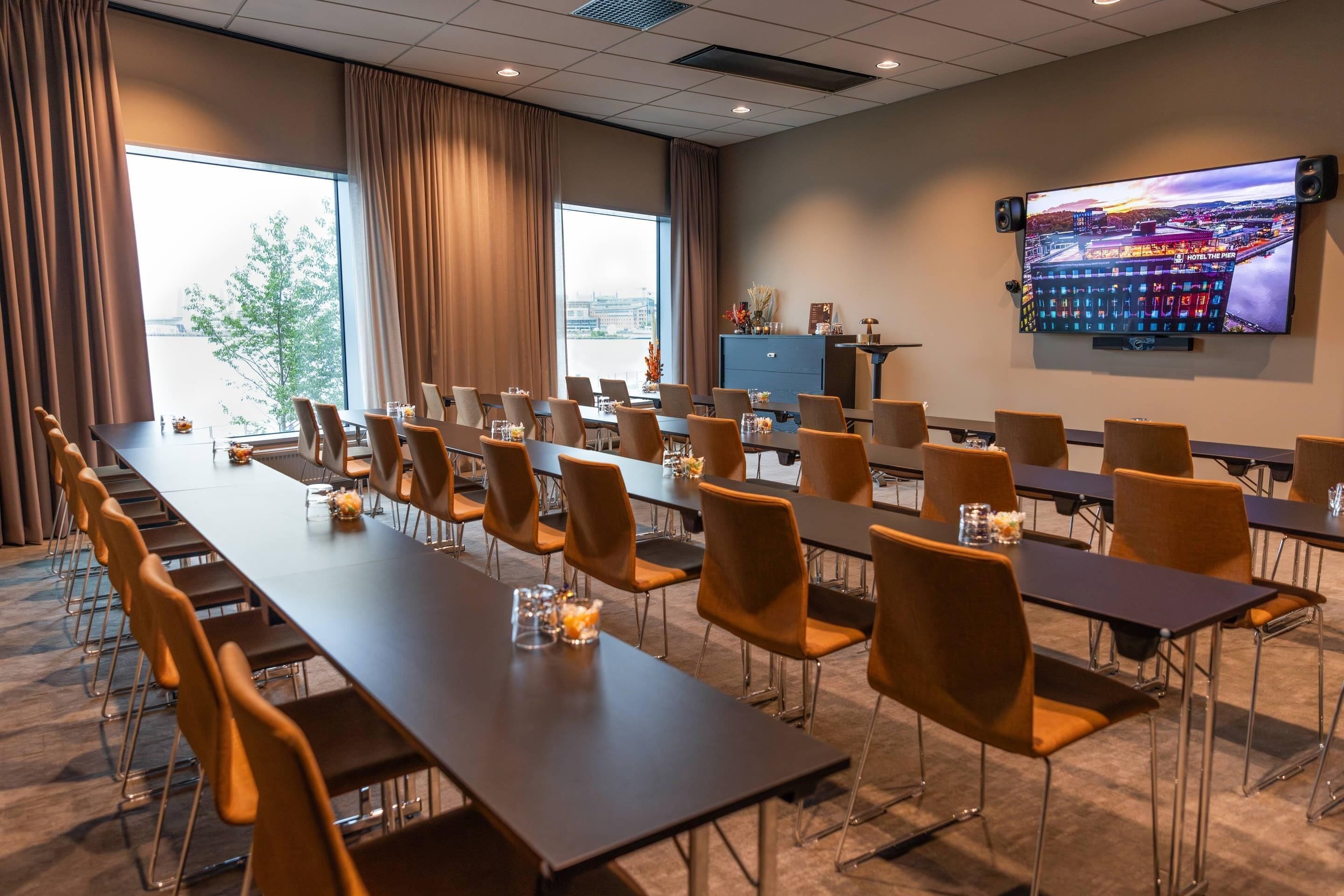 Un salón de conferencias con mesas largas, sillas, una pantalla grande y vistas por las ventanas.