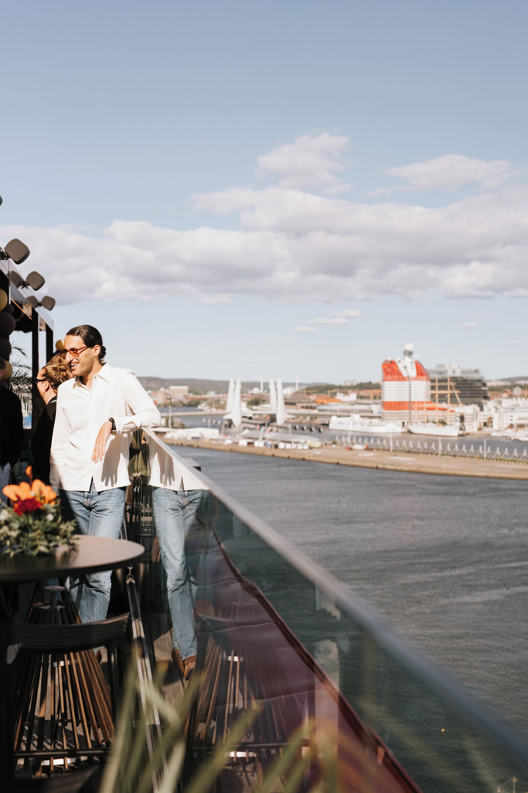 A man standing on a balcony overlooking a body of water with a bridge and industrial structures in the background.