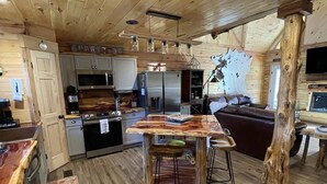 A rustic cabin interior with a wooden floor, a central island table, a sofa, and a television mounted on a wall. - Newly built LOG Cabin-Float, Golf, Hike, Hunt, Fish, visit a winery...Relax! (cuba)