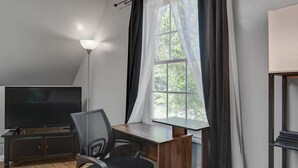 Ein Zimmer mit Flachbildfernseher, Schreibtisch, Stuhl und einem Fenster mit Vorhängen.