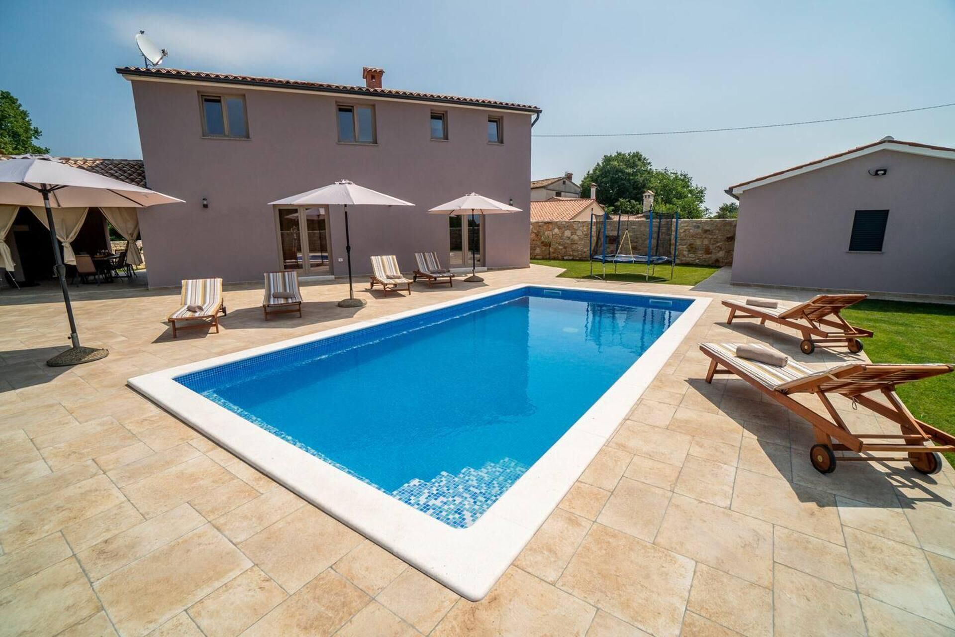 A rectangular swimming pool with lounge chairs and umbrellas, surrounded by a tiled patio and a building with a tiled roof.