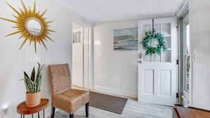A hallway with a sunburst mirror, a wicker chair, a potted plant, a wreath on the door, and a painting on the wall. - Key Lime Cottage - Updated - 1 Minute Walk to Beach & Pier (Flagler Beach)