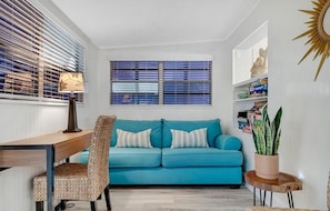Una habitación con un sofá azul, un escritorio de madera, una silla, una ventana con persianas y una planta en maceta.