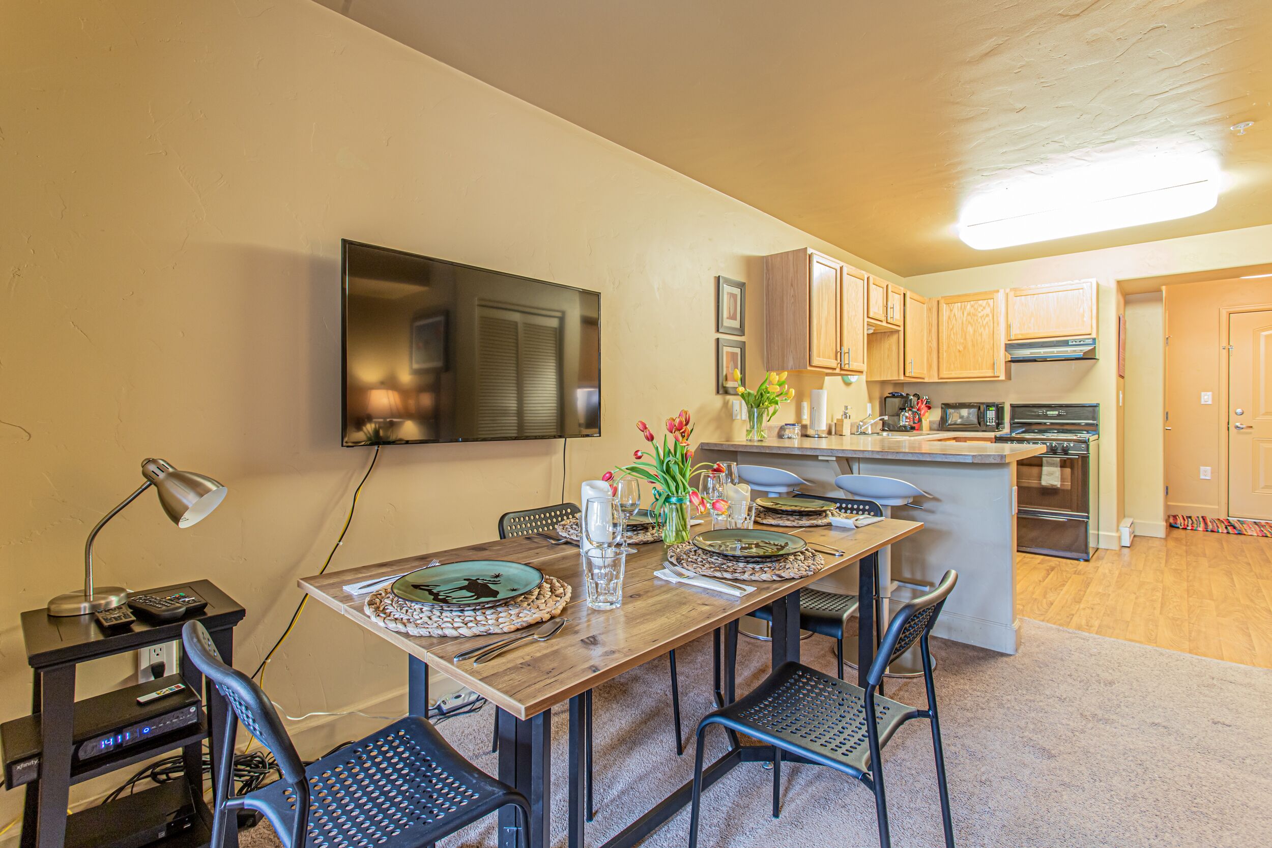 A dining area with a wooden table set for four, a television mounted on the wall, and a kitchen in the background.