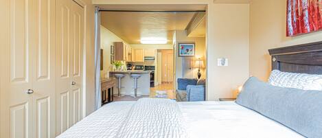 A hotel room with a large bed, a desk, and a kitchenette in the background.