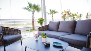 A modern outdoor seating area with a sofa and chairs, a round table with a plant and a bowl, and a view of palm trees and a distant shoreline. - Building Hotel (Carinaro)