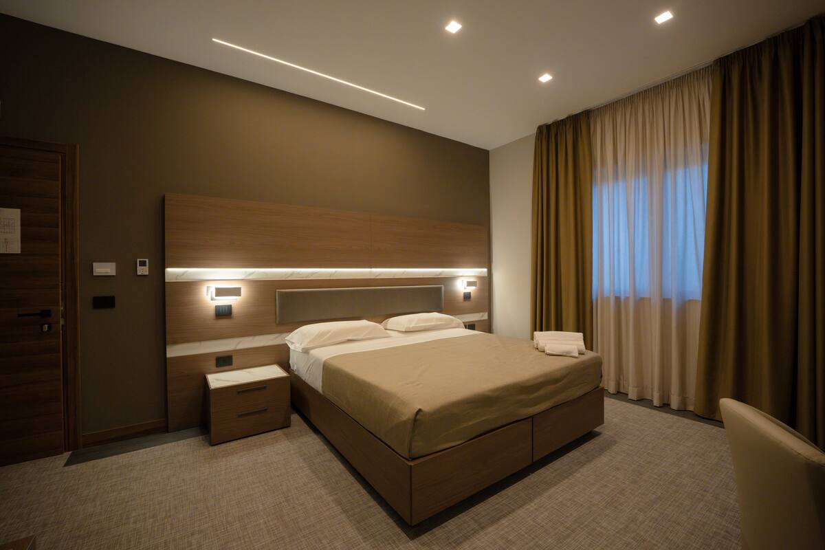 Quarto de hotel moderno com uma cama grande, mesas de cabeceira, um criado-mudo e uma janela com cortinas.