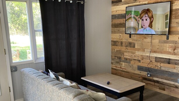 Un salon comprenant un canapé d’angle, une table basse, un téléviseur fixé au mur en bois et un ventilateur de plafond.