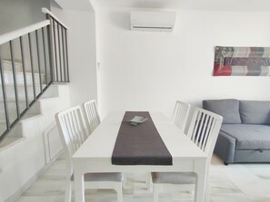 Een witte eettafel met een donkere tafelkleed, witte stoelen, een grijze bank en een trap met een metalen leuning.