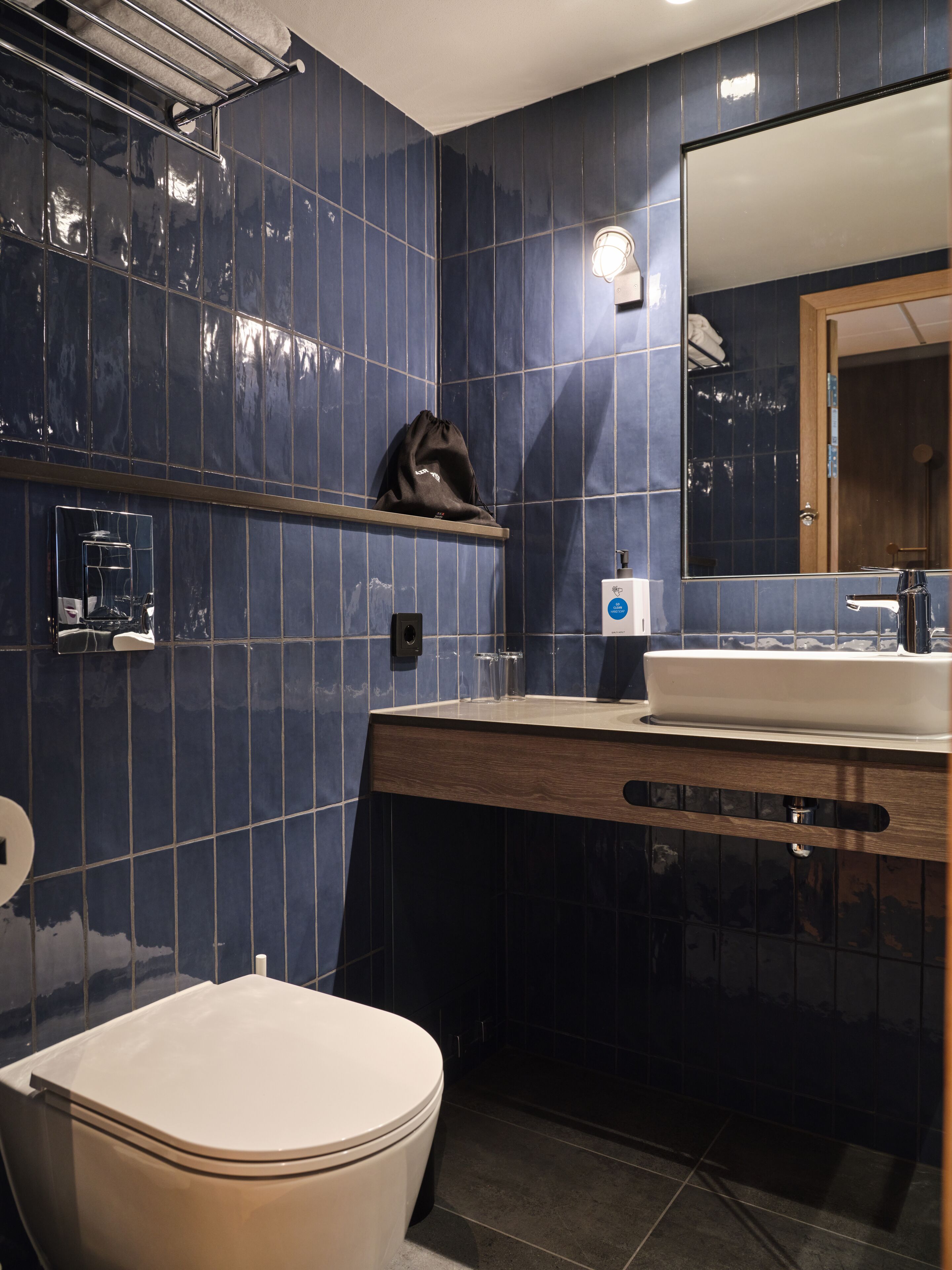 Un baño moderno con azulejos azul oscuro, un inodoro blanco, un tocador de madera con un espejo y un perchero para toallas.