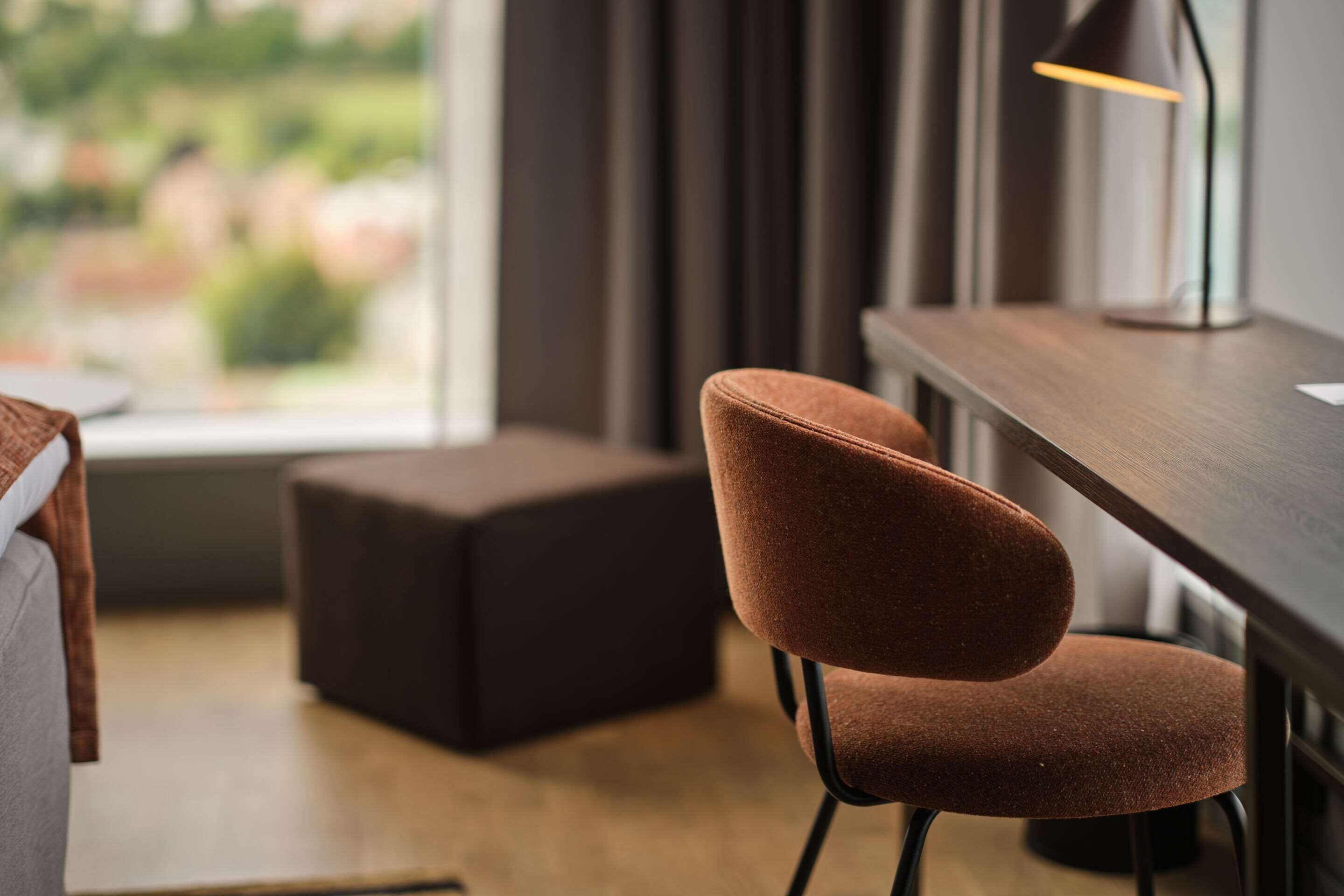 Una habitación moderna con un escritorio de madera, una silla tapizada en tonos marrones, un pequeño reposapiés y vistas a zonas verdes a través de una ventana.