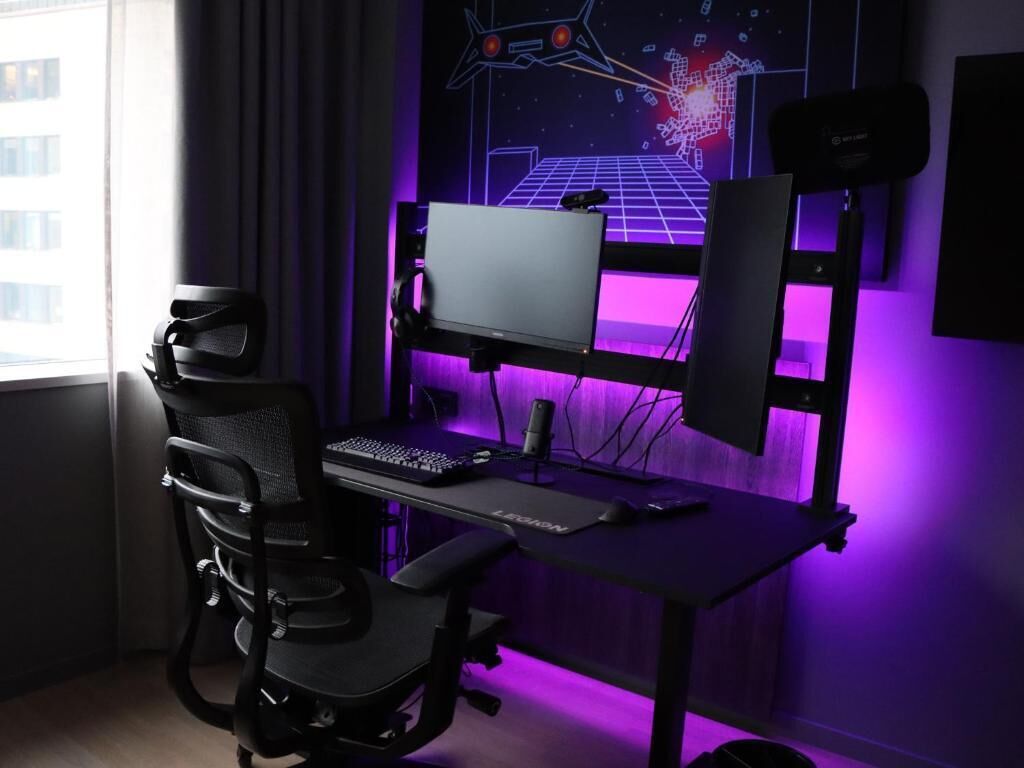 A gaming setup with a monitor, keyboard, and chair in a room with purple lighting and a digital art wall.