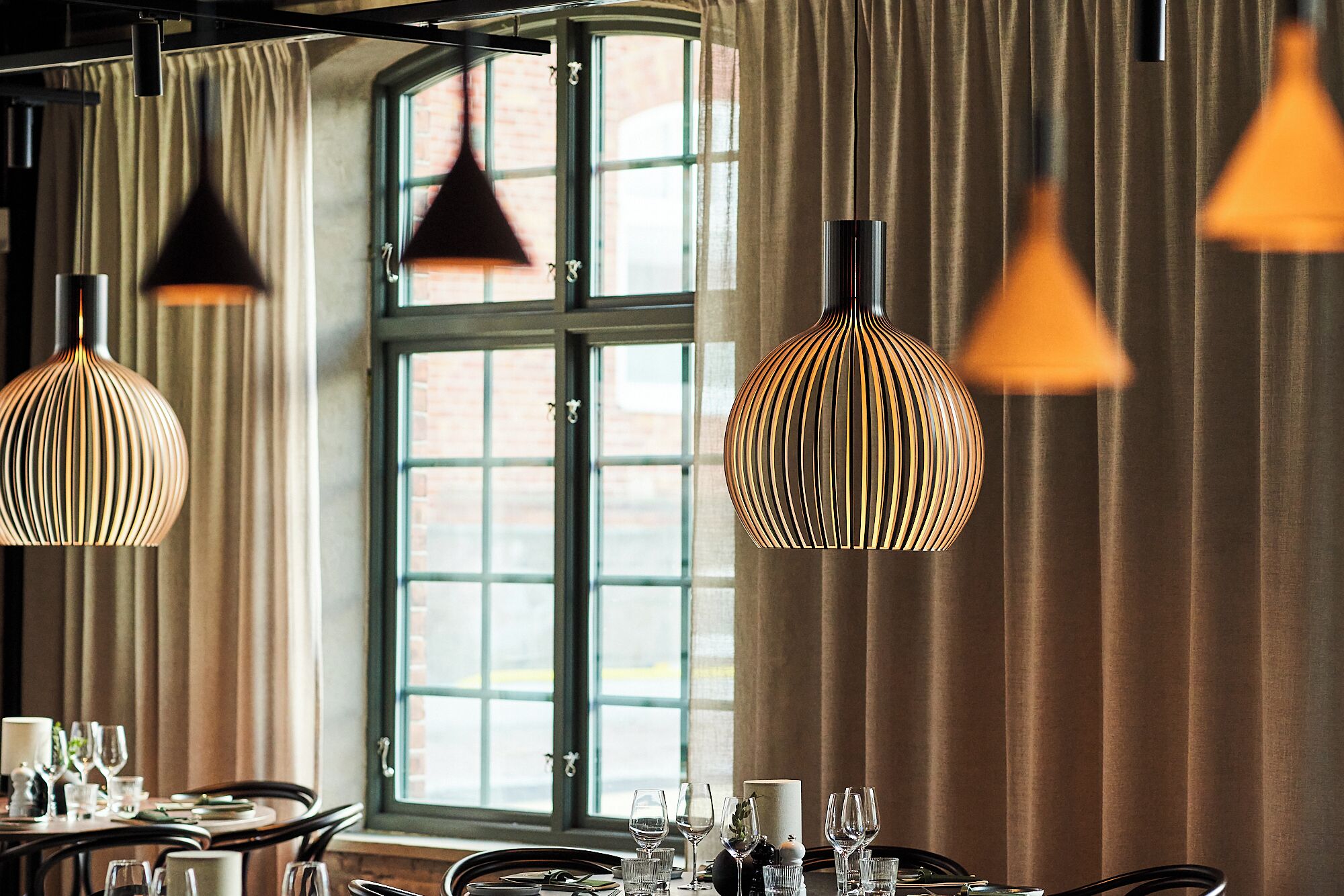 A dining area with a table set for a meal, a window with curtains, and pendant lights.