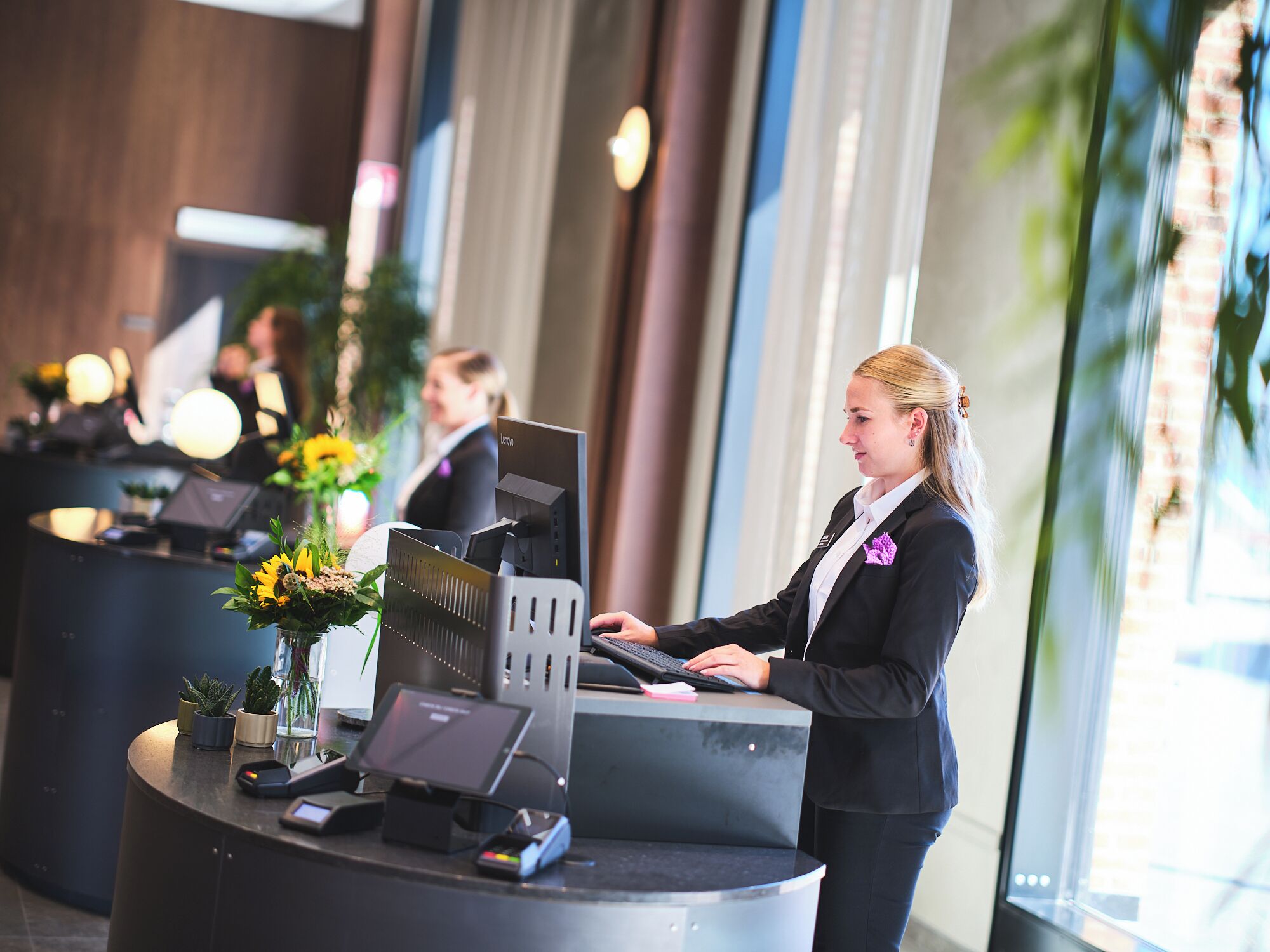 A reception desk with a computer, a cash register, and a credit card reader. There are potted plants on the counter.