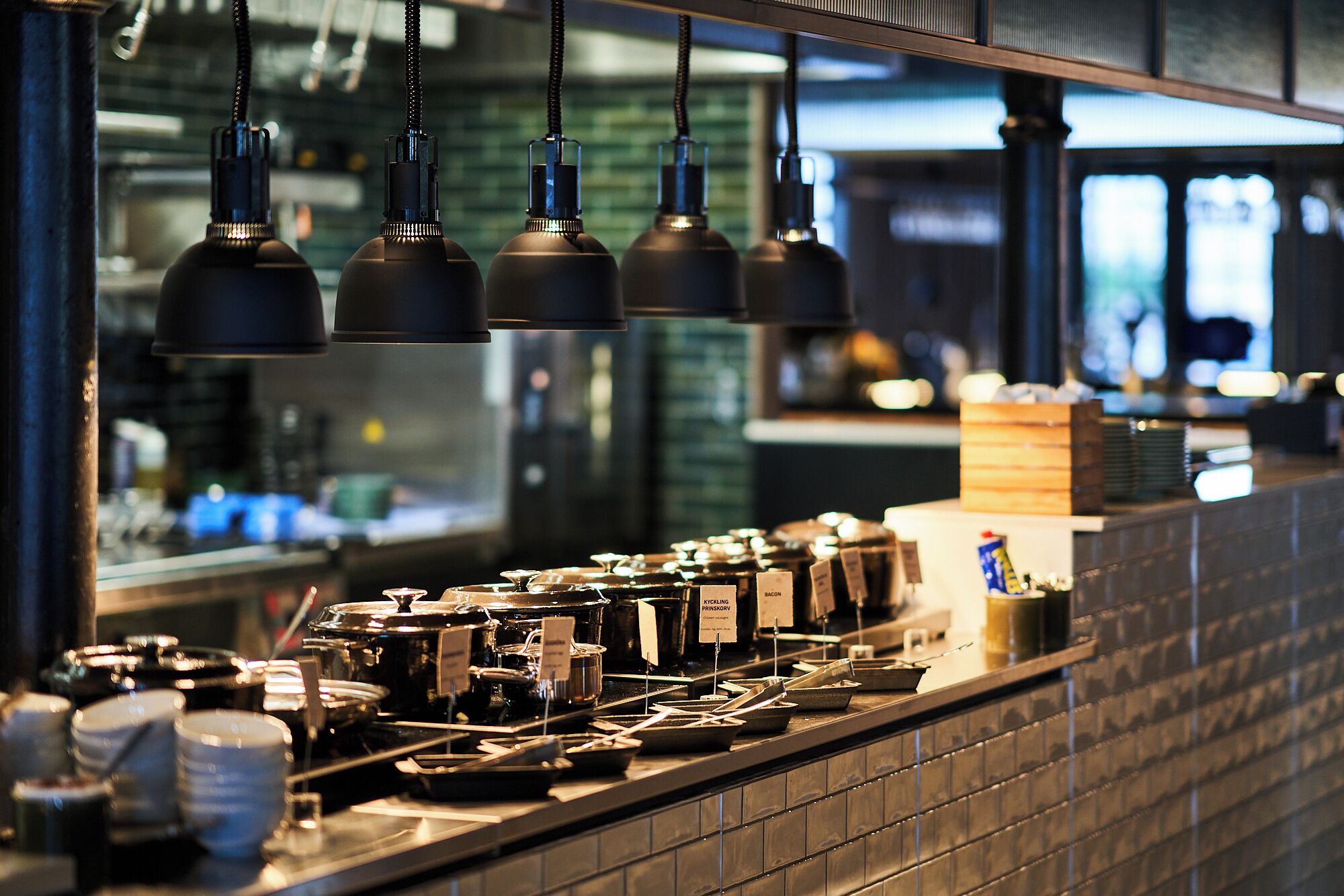 A kitchen counter with various cooking utensils and containers, a window with a view of the city, and hanging kitchen lights.